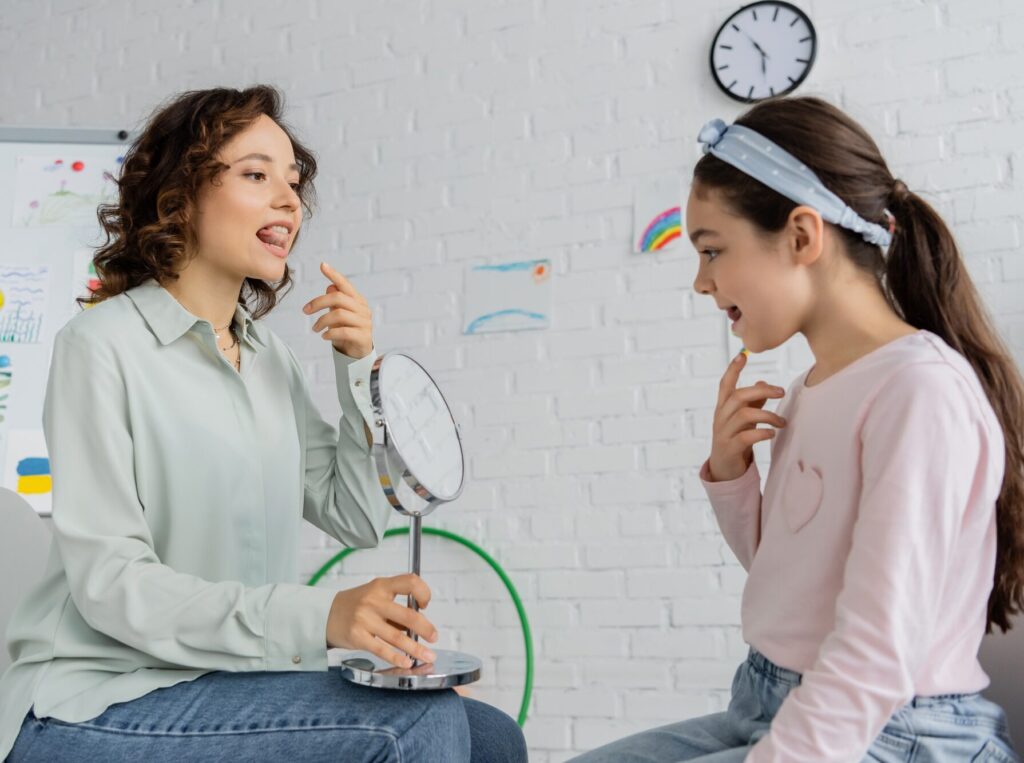 Speech therapist holding mirror and pointing at mouth near pupil in consulting room